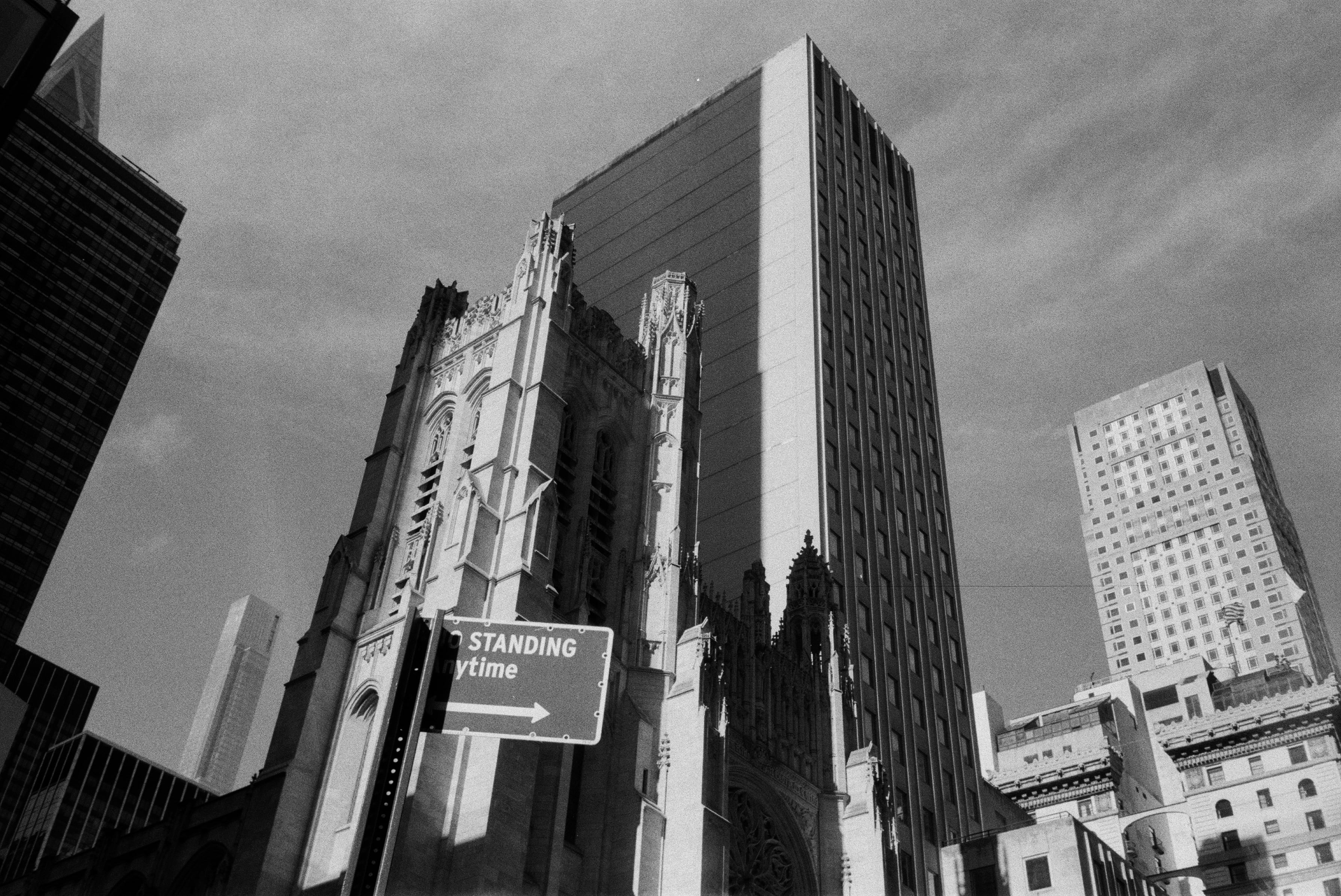 A black and white photo of a New York City landscape. The subject of the photo is a historic catherdral that is having hard light cast on it by a modern skyscraper that towers behind it.
